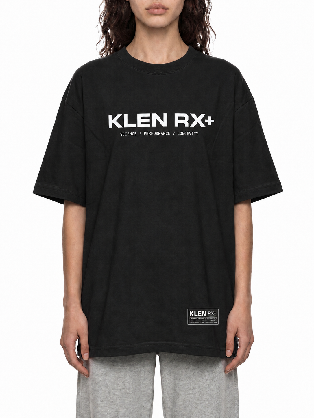 A model wearing an oversized black Klen RX+ t-shirt with white text on the chest and a small white Klen patch near the bottom hem, paired with light gray sweatpants against a white background.